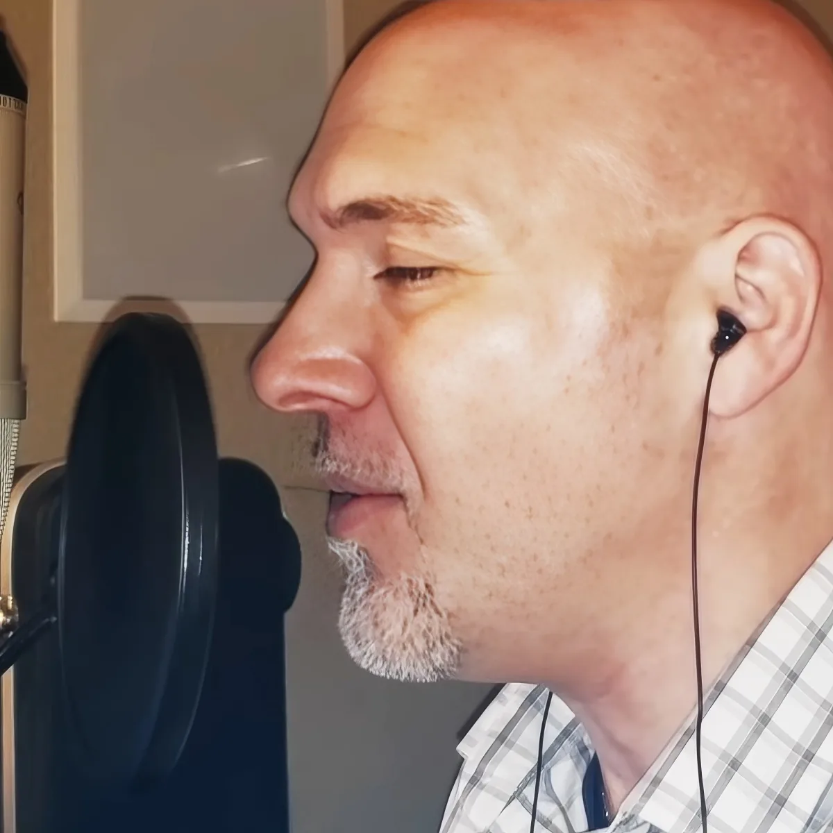 A bald man with earphones sings into a microphone in a recording studio.