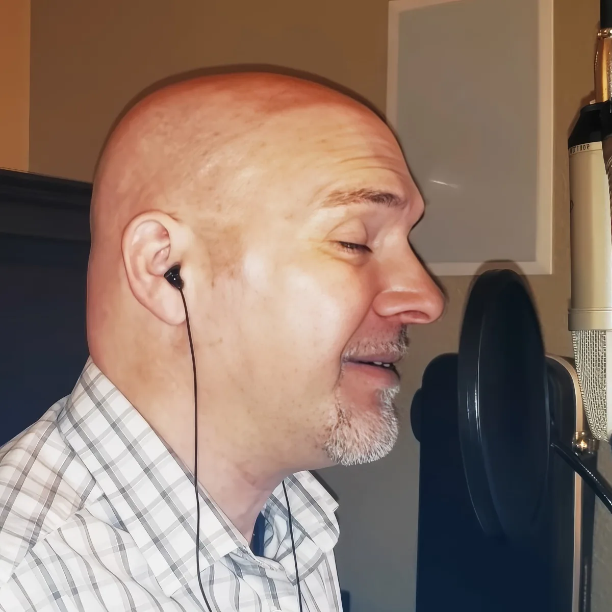 A man with a bald head and earphones smiles while speaking into a microphone in a recording studio.