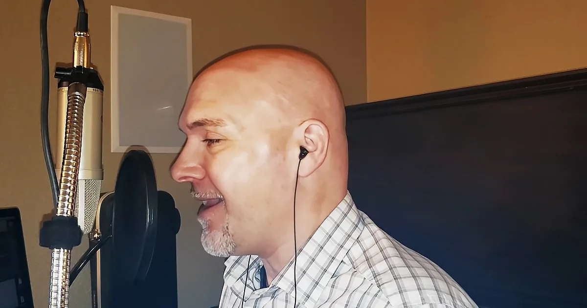 A man in a plaid shirt speaks into a microphone in a recording studio.