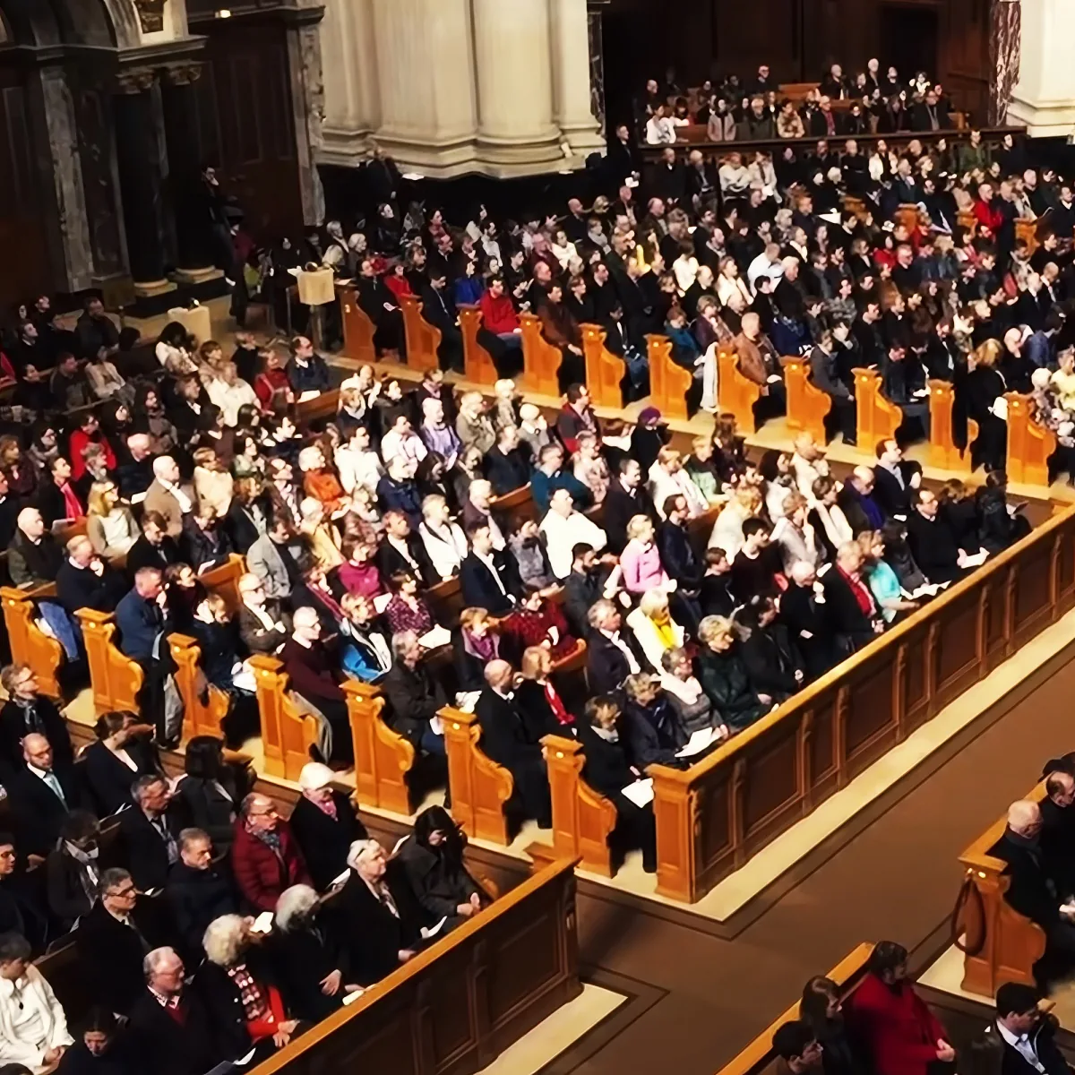 A large crowd of people is seated in rows of wooden pews in a grand hall.