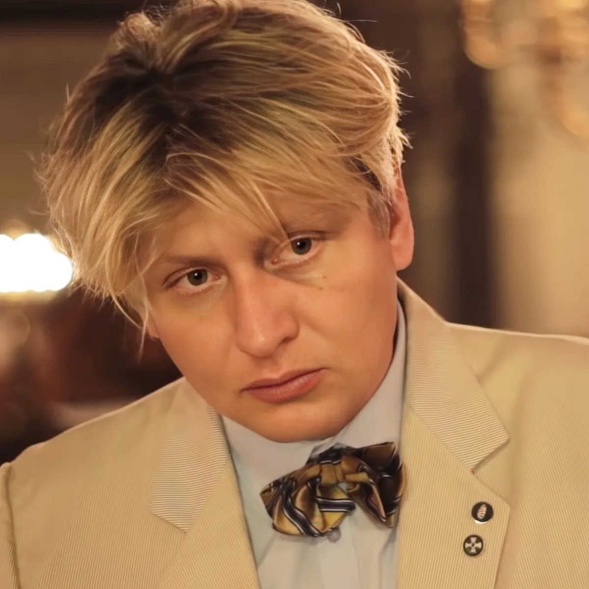 A person with blonde hair, wearing a beige suit and a patterned bow tie, is looking to the side.