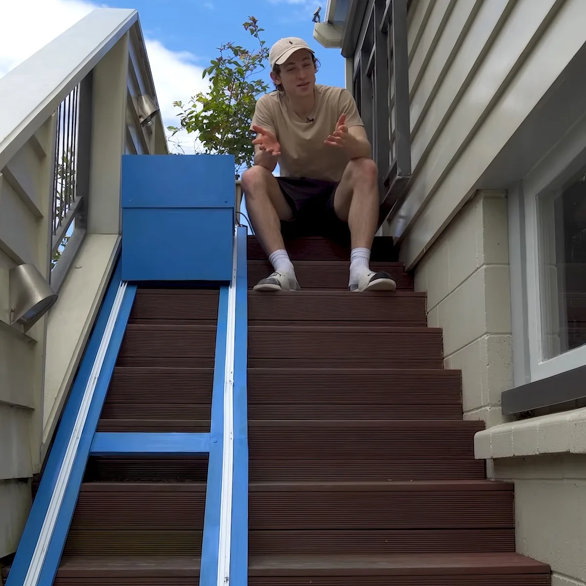 A person sits on a brown staircase with a blue handrail, wearing a beige shirt and cap, gesturing with their hands.