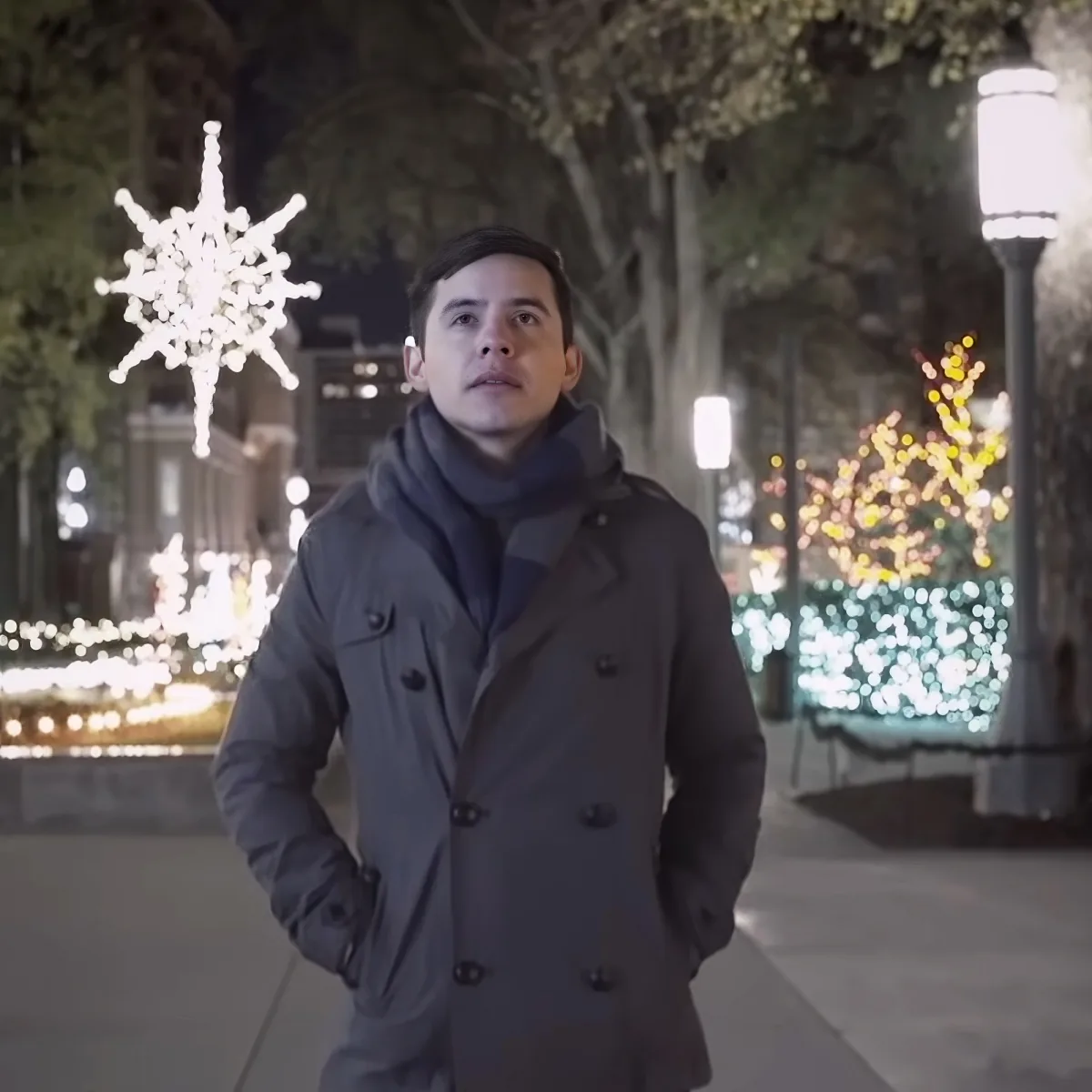 A man in a gray coat stands on a sidewalk, gazing upward, surrounded by festive holiday lights.