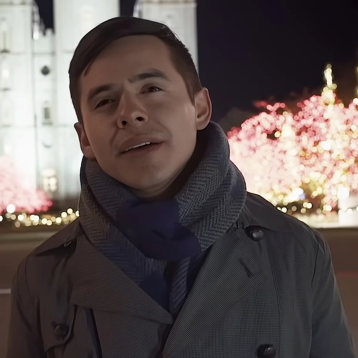 A man in a trench coat and scarf stands in front of a festive, illuminated Christmas tree.