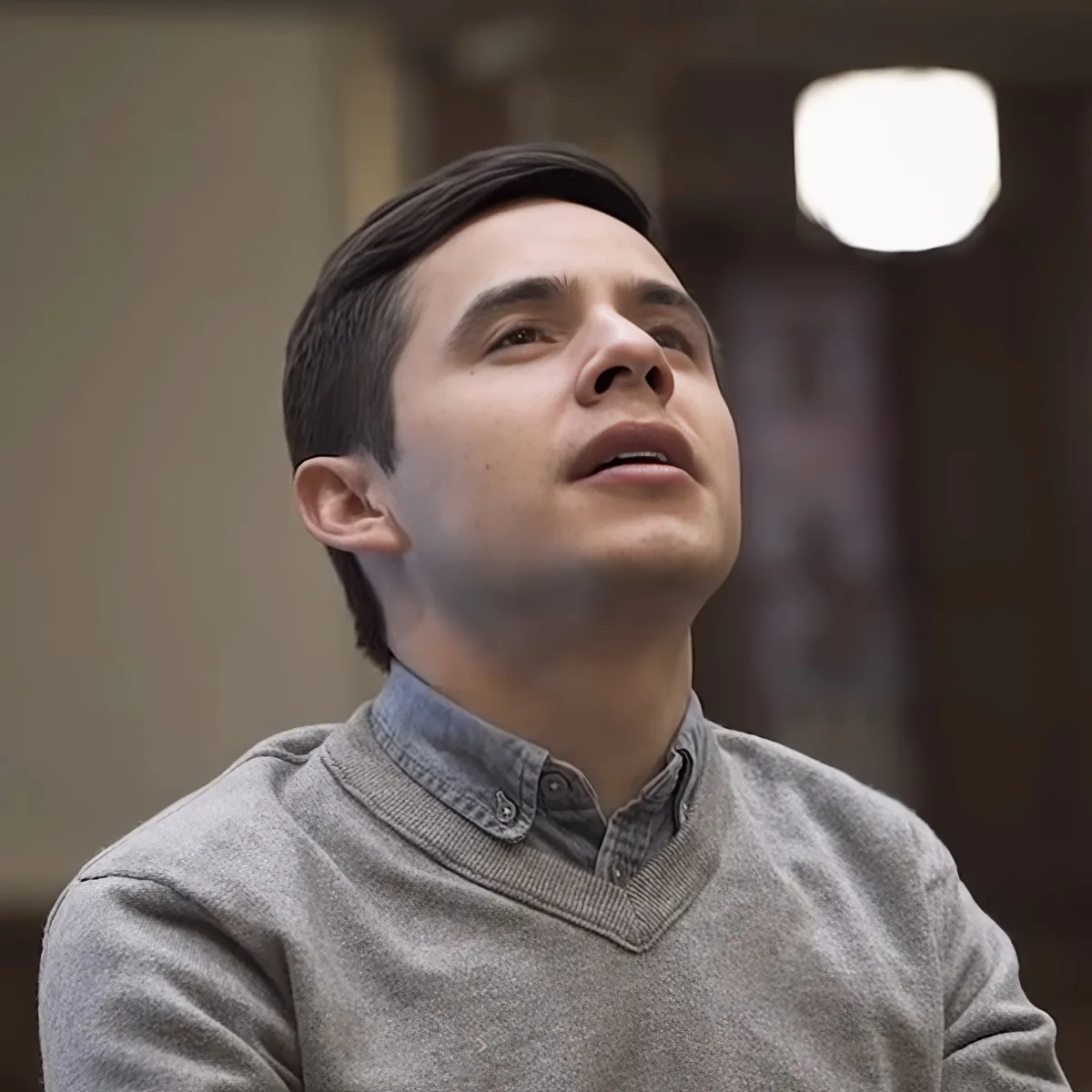 A person in a gray sweater and button-up shirt looks upward, mouth slightly open, in a softly lit indoor setting.