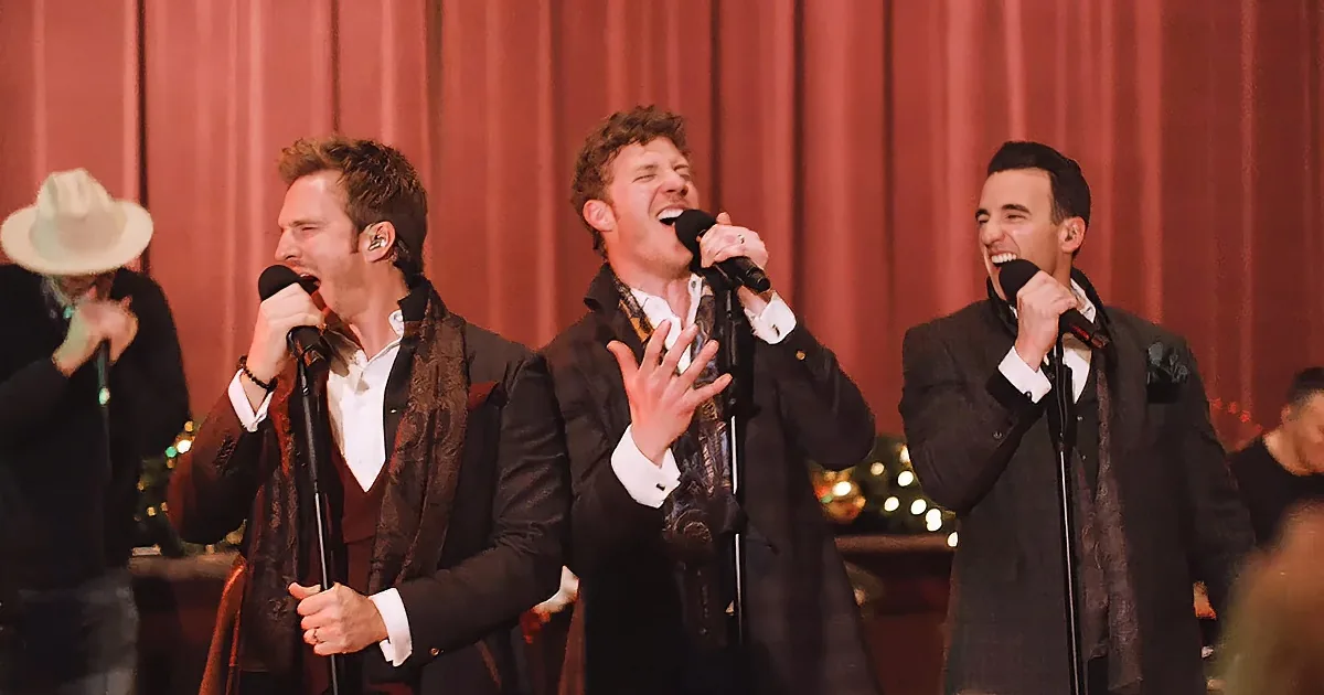 Three men in suits sing into microphones on stage, one in a cowboy hat in the background.