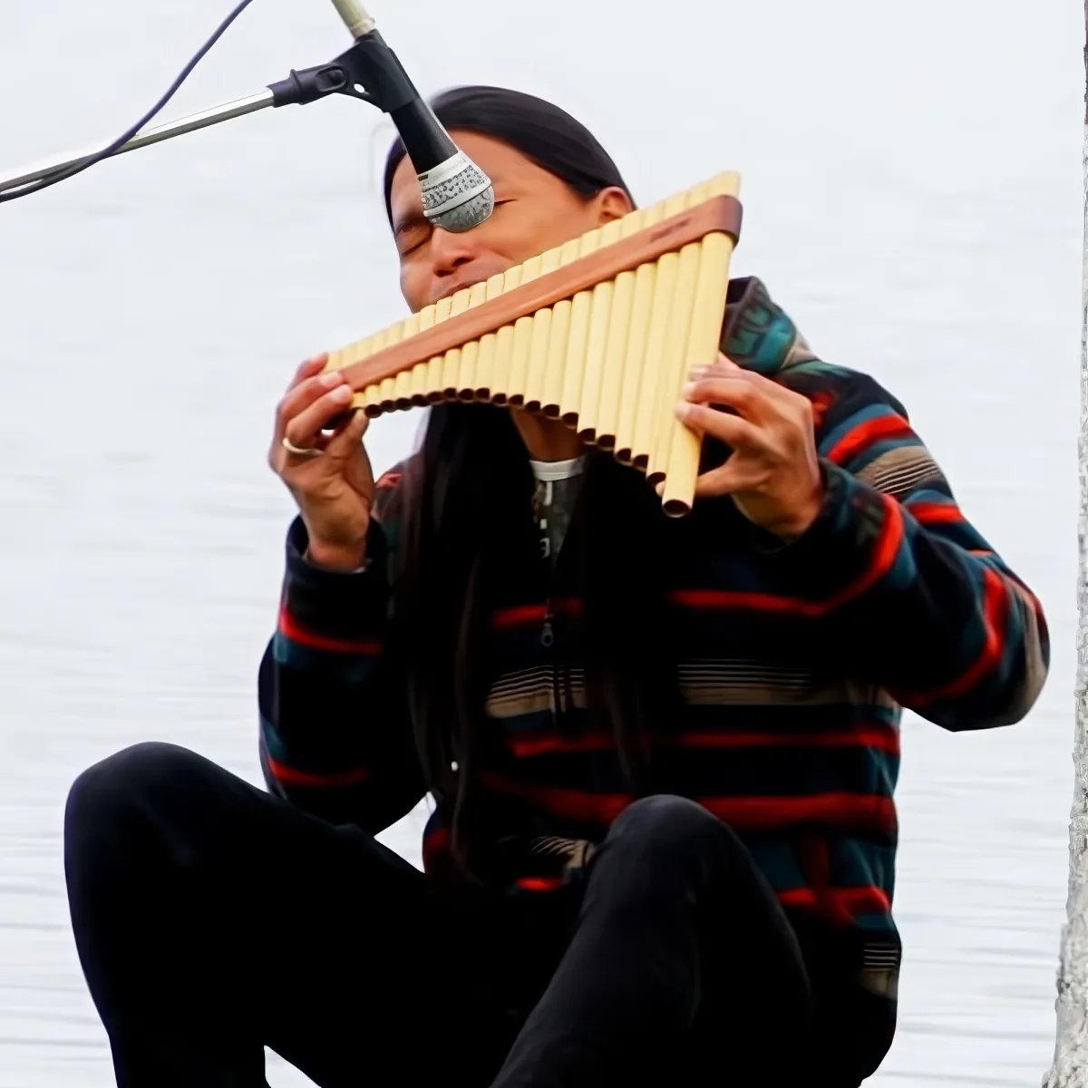 A person in a striped sweater plays a pan flute near a microphone, with a body of water in the background.