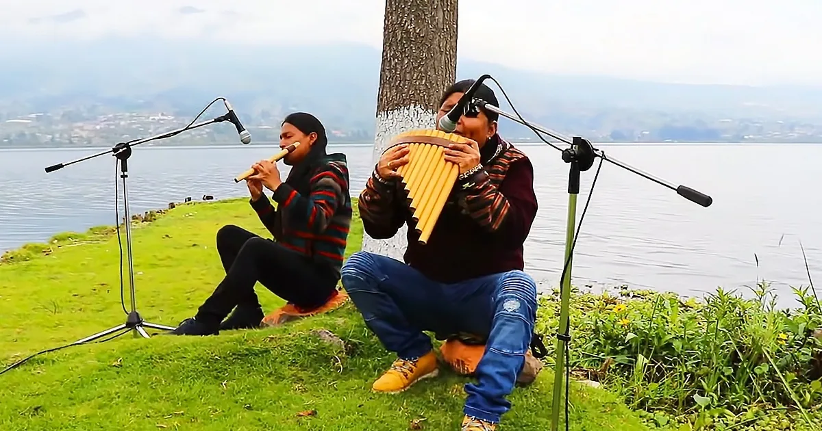 Two musicians play flutes and a pan flute by a lake, with microphones and a scenic backdrop.