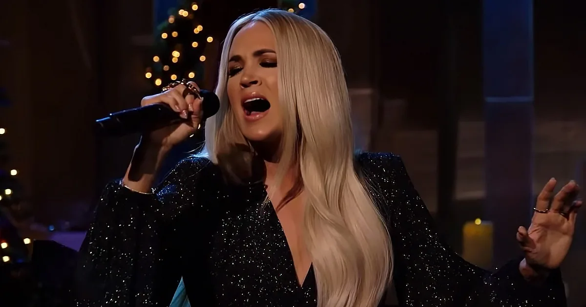 A woman with long blonde hair sings into a microphone, wearing a sparkly black dress, with a Christmas tree in the...