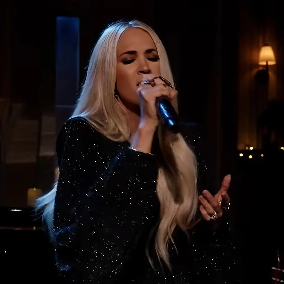 A woman with long blonde hair sings into a microphone, wearing a black sequined top.