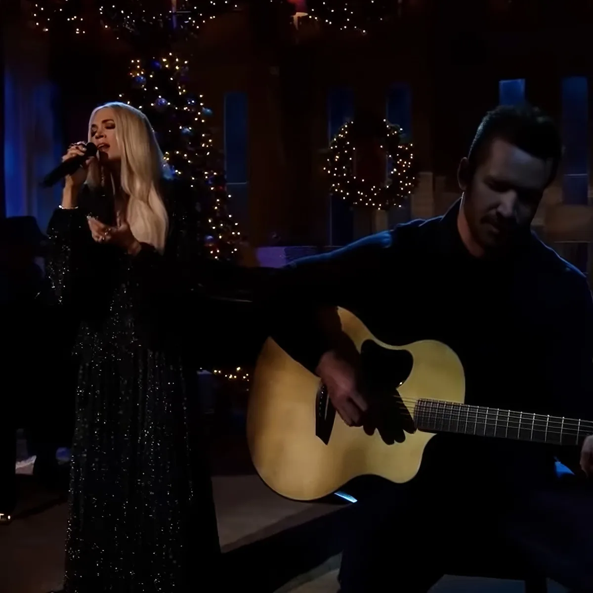 A woman sings into a microphone, a man plays guitar, both in a festive setting with Christmas decorations.