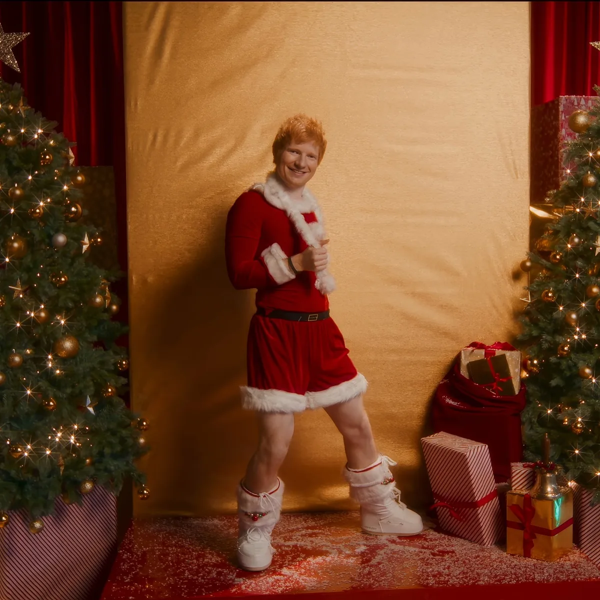 A person in a festive Santa outfit stands between Christmas trees and presents, smiling.