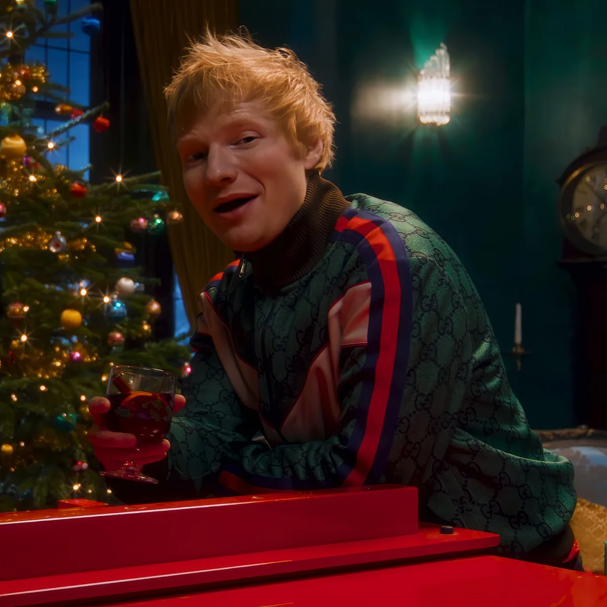 A person with red hair, wearing a green Gucci sweater, holds a drink, sitting in front of a Christmas tree.