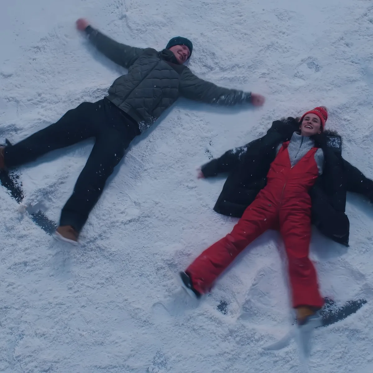 Two people making snow angels in the snow, dressed warmly, enjoying the winter scene.