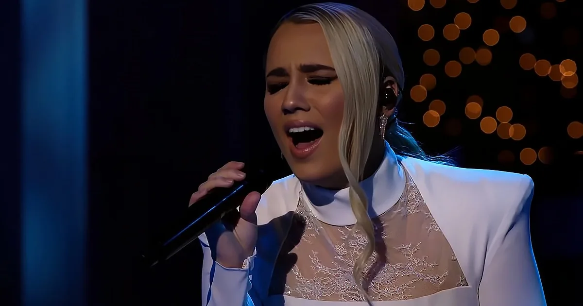 A person with long blonde hair, wearing a white outfit, sings passionately into a microphone on stage.