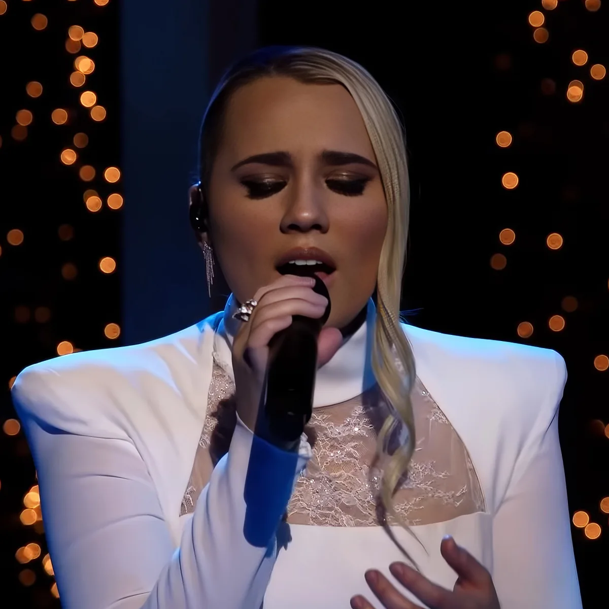A person in a white outfit sings into a microphone, with a blurred background of warm lights.