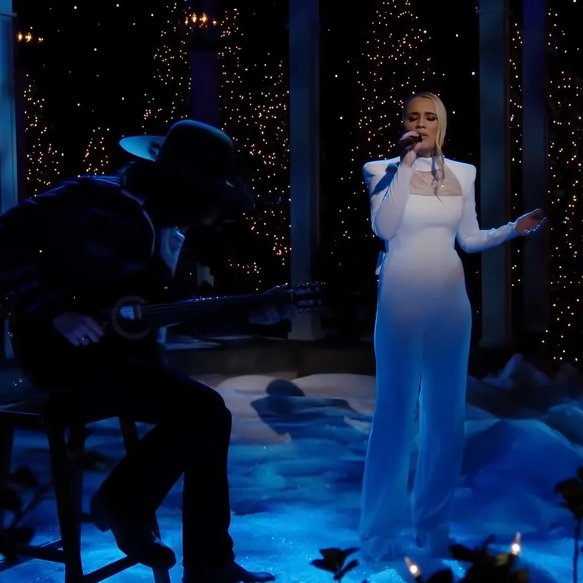 A pregnant woman in a white gown sings on stage, accompanied by a guitarist in a hat.