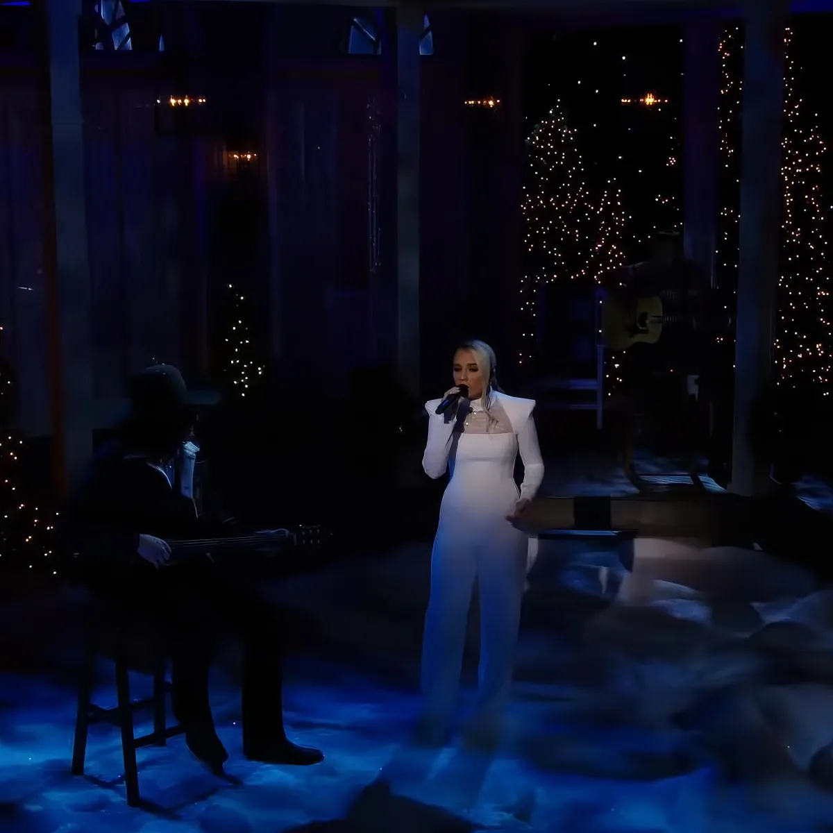 A woman in a white outfit sings into a microphone on a stage with a guitarist and Christmas lights in the background.