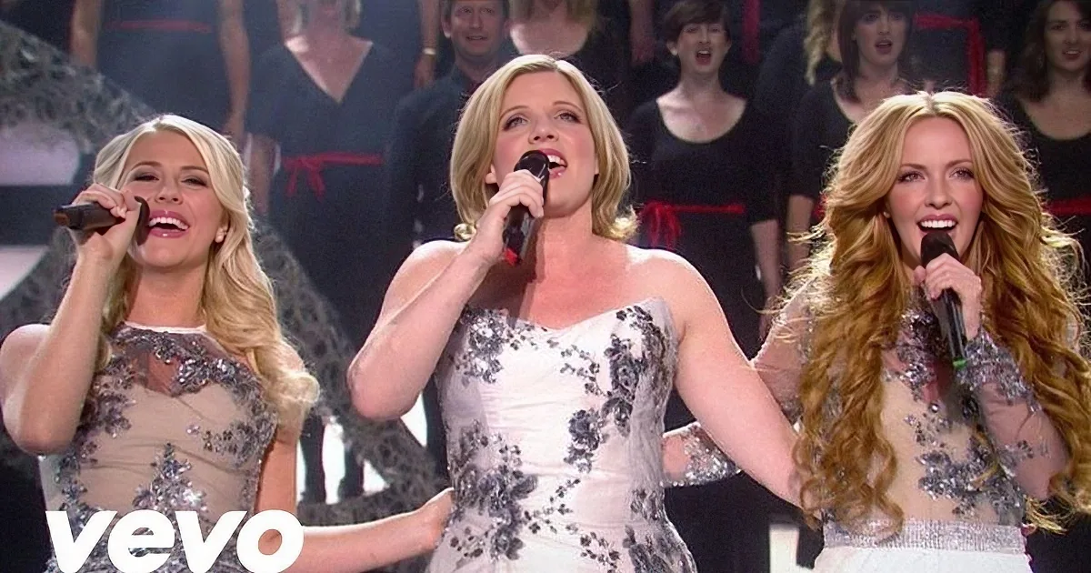 Three women in elegant white dresses sing on stage with a choir and Christmas decorations.