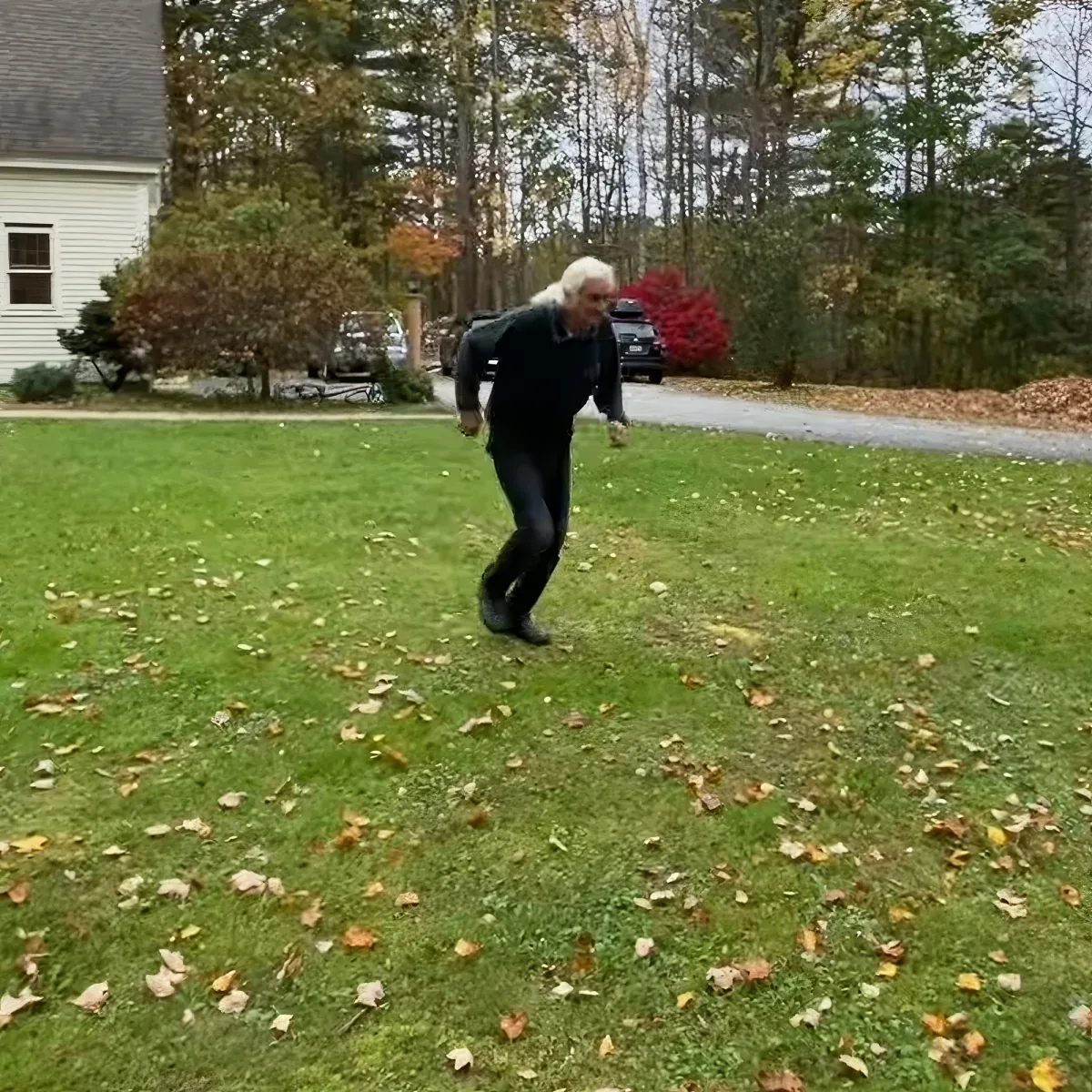 An older man in black runs across a grassy yard with scattered leaves, surrounded by trees and a house.