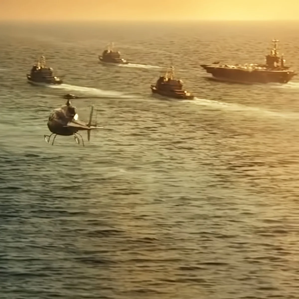 A helicopter flies over the ocean, with four ships in the background, creating a trail of water spray.