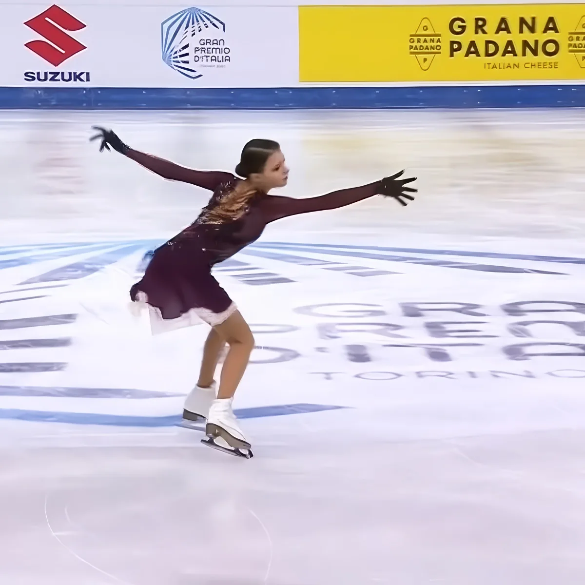 A figure skater in a maroon dress glides gracefully on ice, arms outstretched, at the Gran Premio d'Italia in Turin.