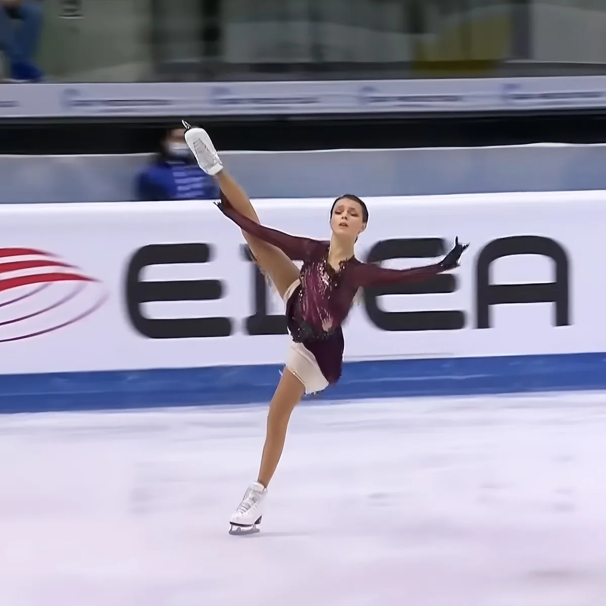 A figure skater in a maroon outfit performs a graceful pose on the ice.