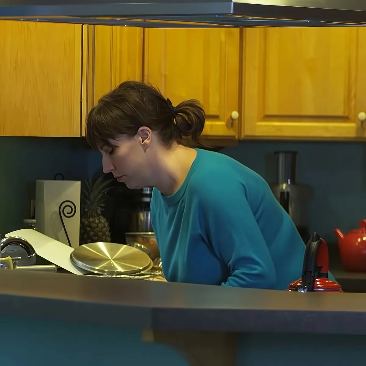 A woman in a teal shirt leans over a kitchen counter, looking down at something.