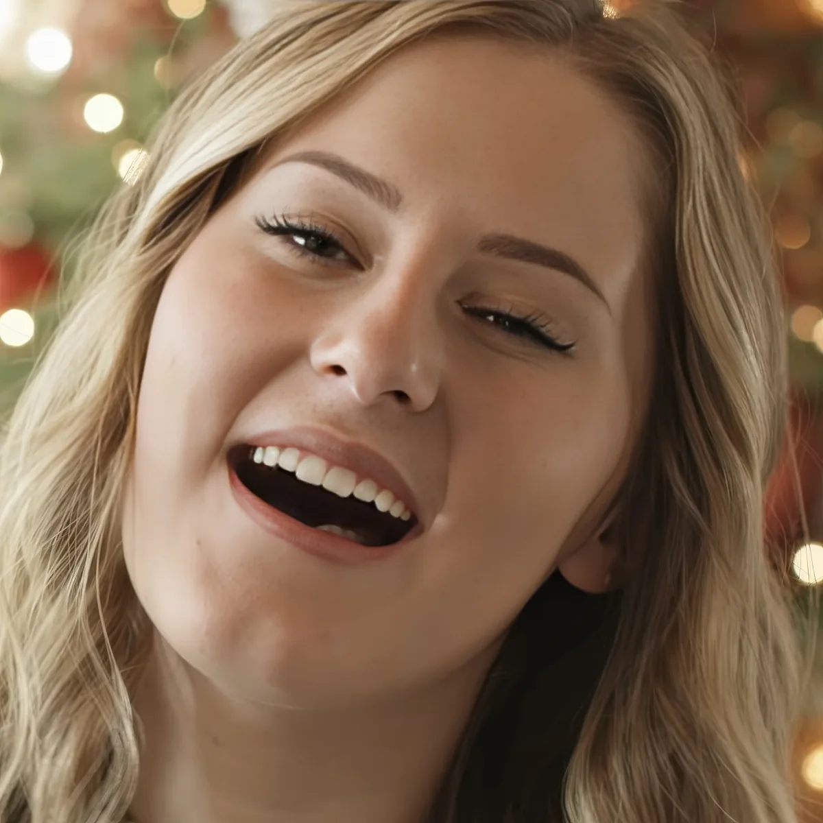 A woman with blonde hair smiles widely, teeth visible, against a blurred Christmas tree backdrop.