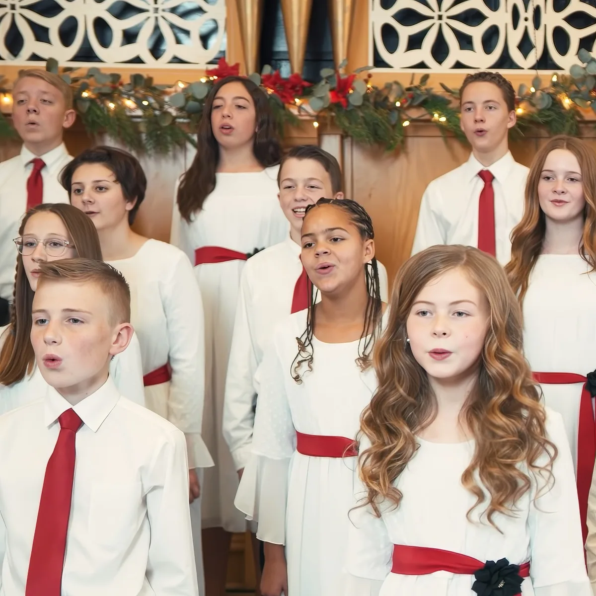 A group of children in white and red choir outfits sing in front of a festive background.