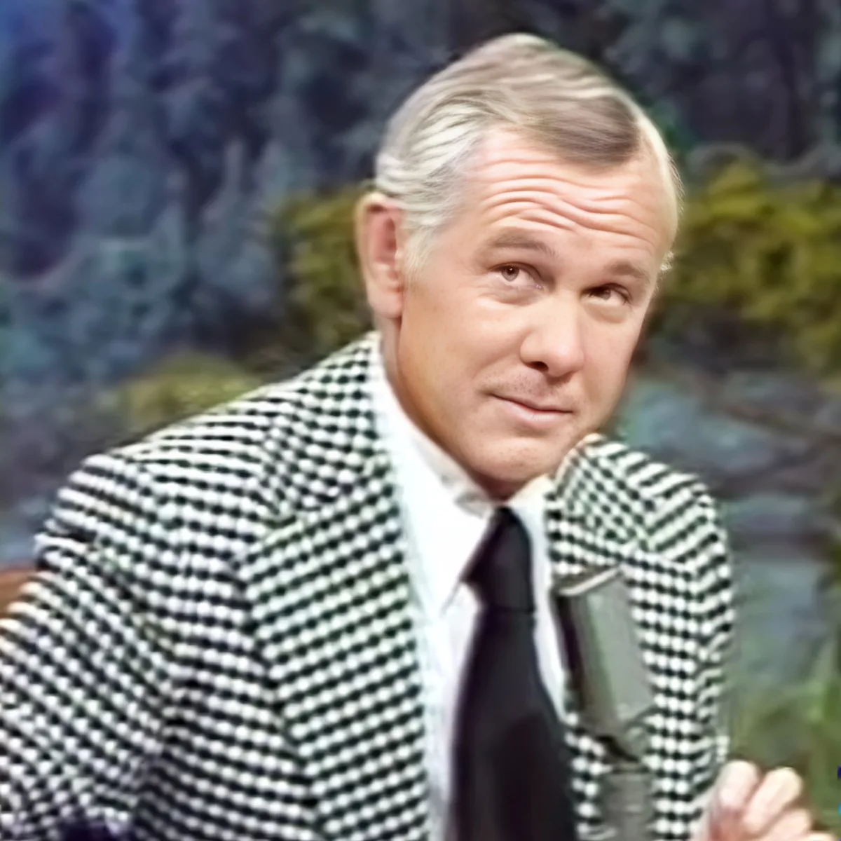 A man in a checkered suit and tie, holding a microphone, appears to be speaking or presenting.