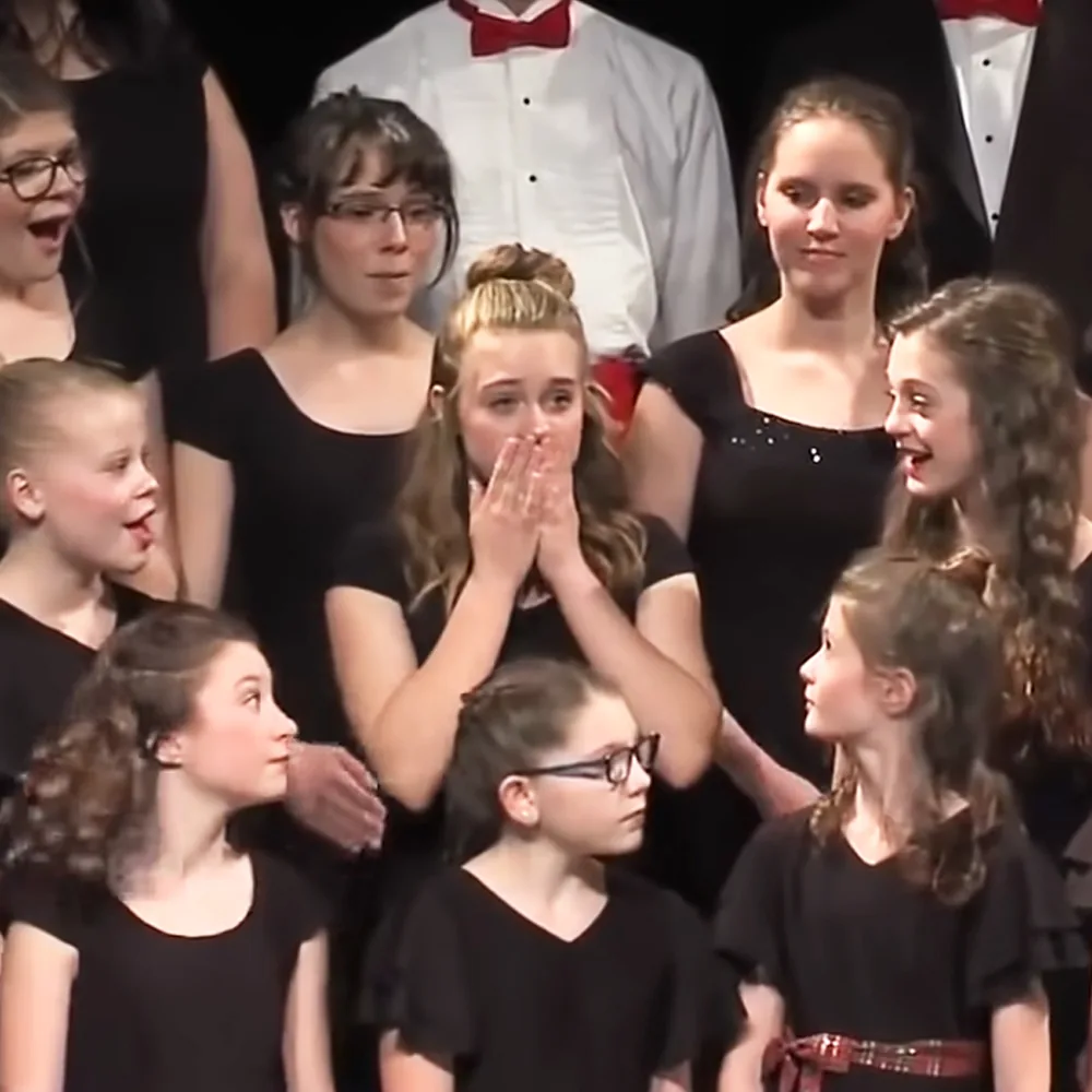 A young girl in a black dress claps her hands while surrounded by other children in a choir setting.