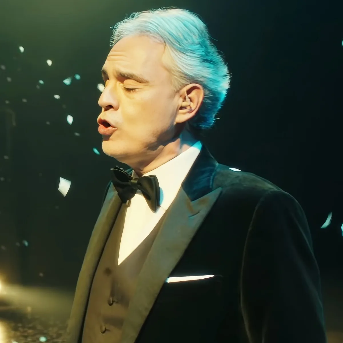 A man in a formal suit with a bow tie and white shirt, possibly singing or speaking on stage.