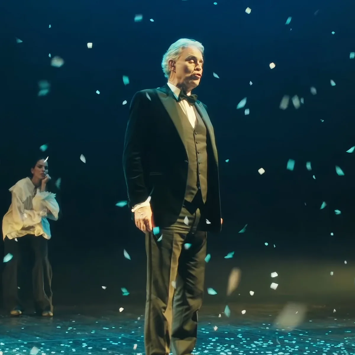 An older man in a suit stands on stage with confetti falling around him, while a woman in a white dress is in the background.