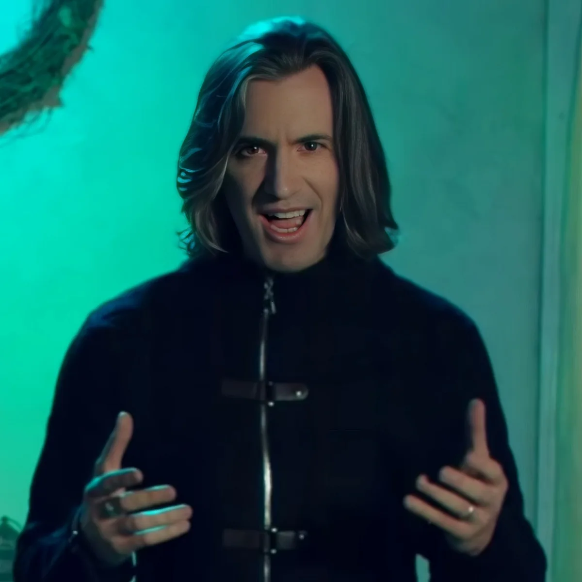A person with long hair and a black outfit is gesturing with their hands in front of a teal background.