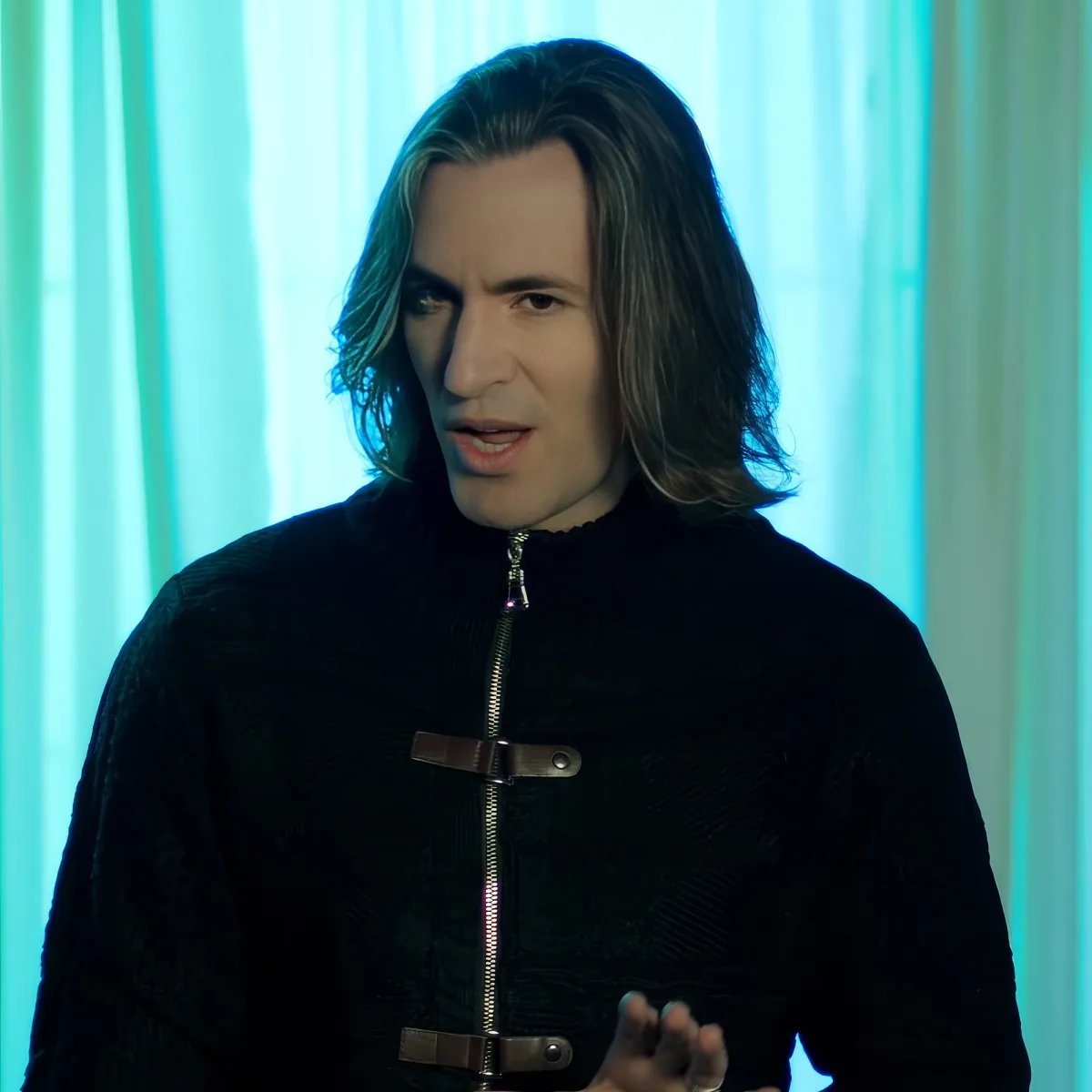 A person with long hair, wearing a black jacket with a zipper and belt, stands against a blue-lit background.