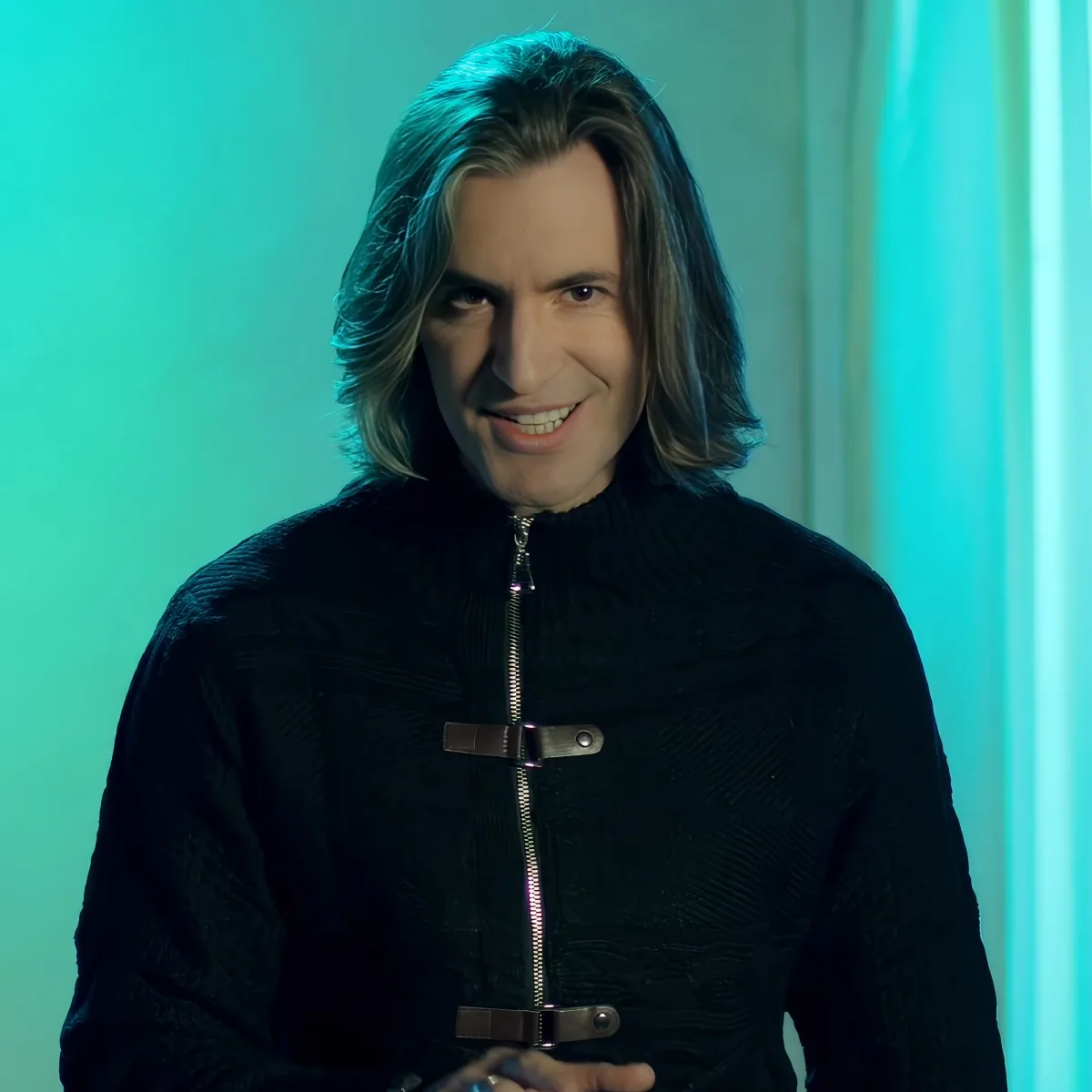 A person with long hair, wearing a black textured jacket with a zipper and belt, smiles against a teal-lit background.