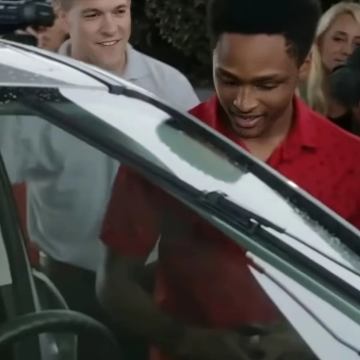 A man in a red shirt is getting into a car, smiling, with a person in a white shirt behind him.