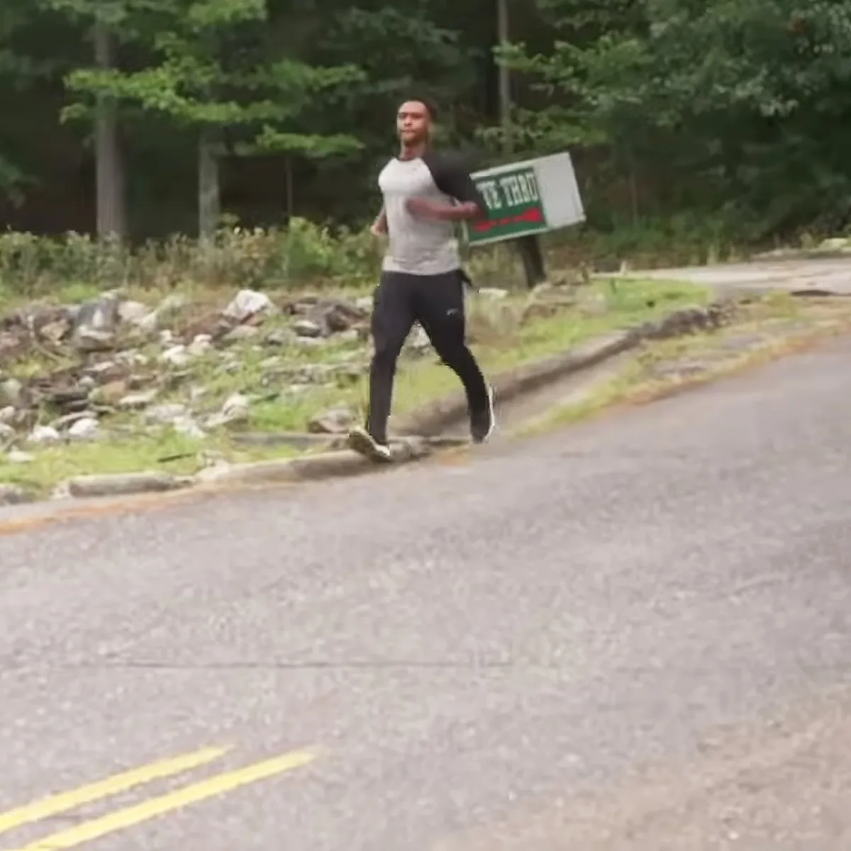 A man in a white tank top and black pants runs on a road near a sign with green and red text.