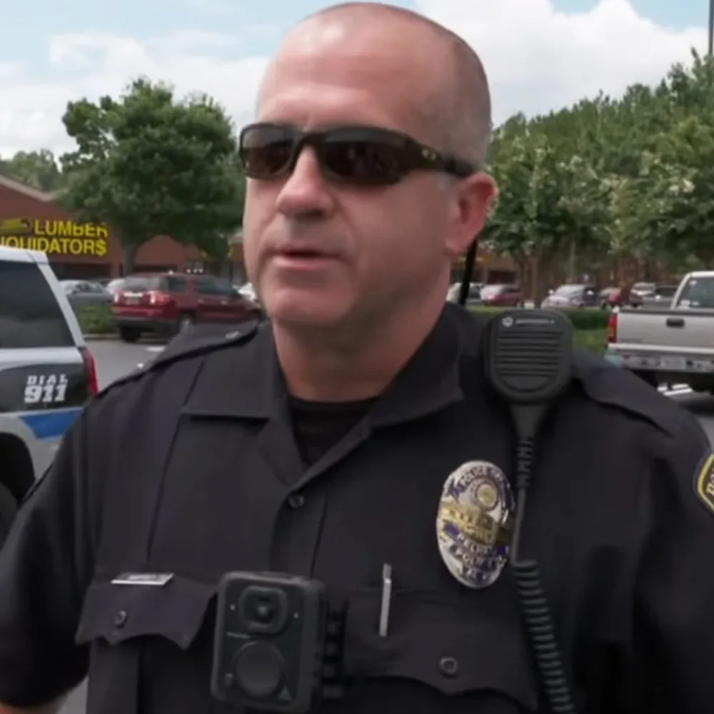 A police officer in uniform stands outdoors, wearing sunglasses, with a badge and radio visible on his chest.
