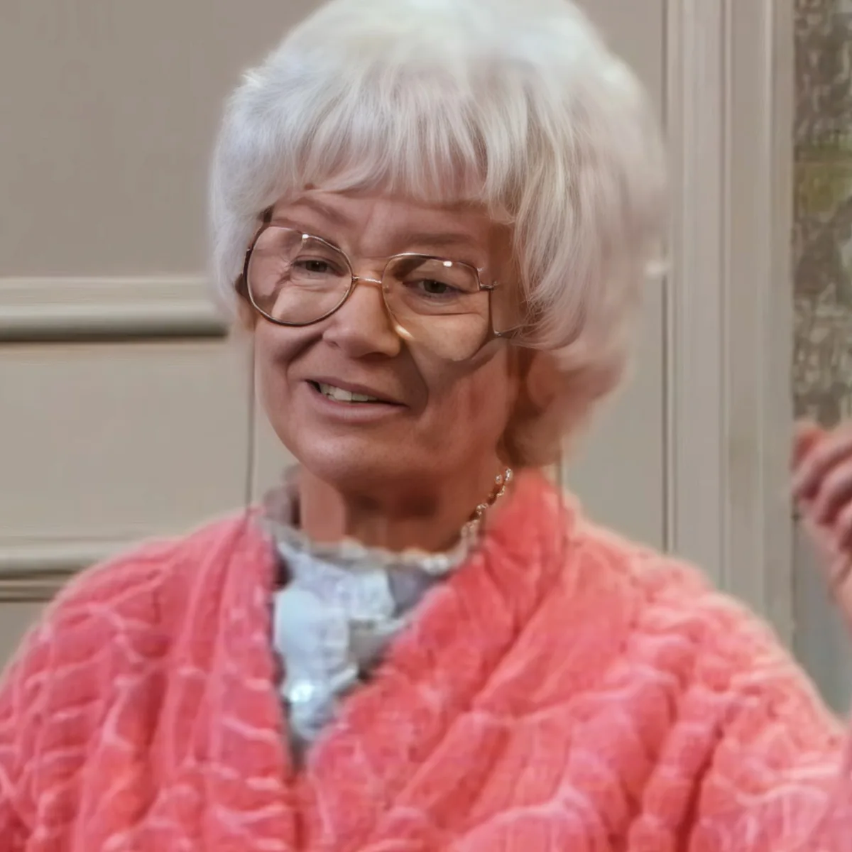 An elderly woman with short white hair, wearing glasses and a pink quilted jacket, smiles warmly.