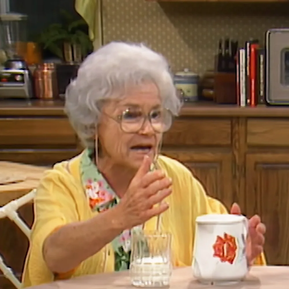 An elderly woman in a yellow cardigan sits at a table, holding a mug and gesturing with her other hand.