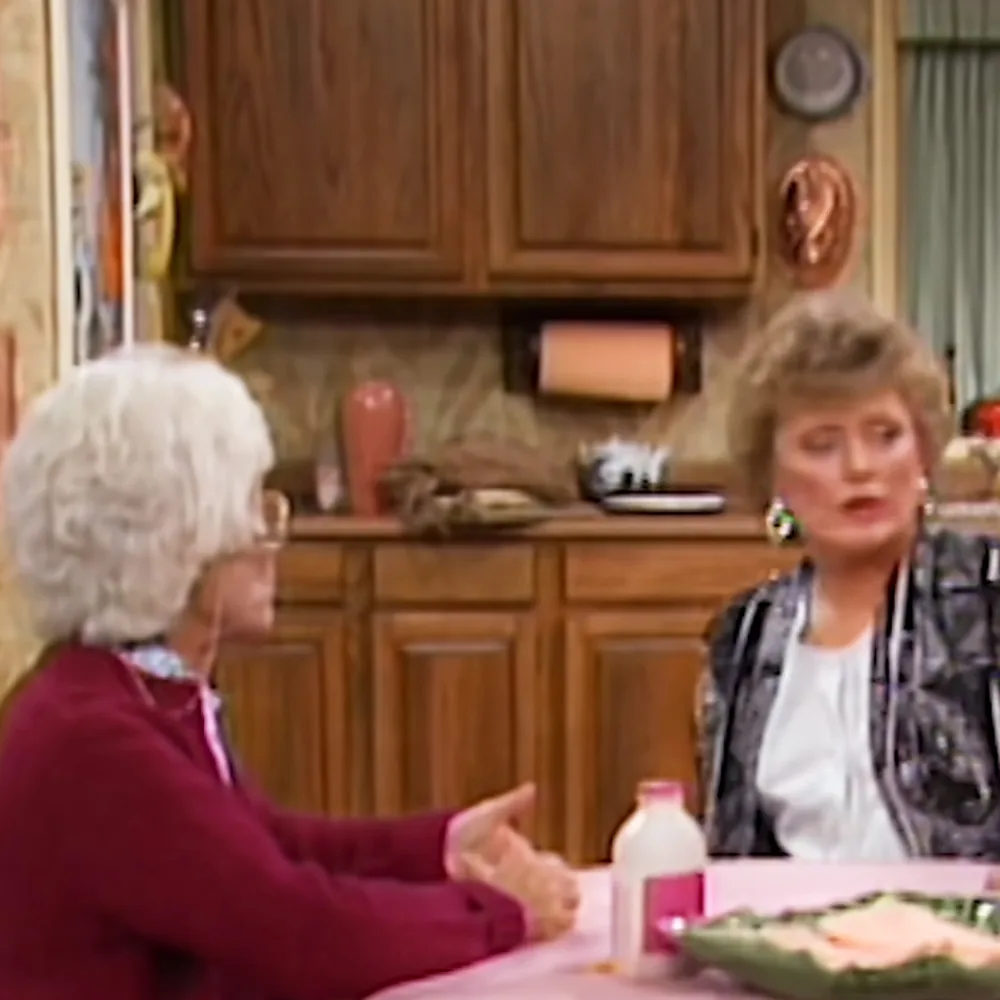 Two women are having a conversation at a kitchen table with food and milk.