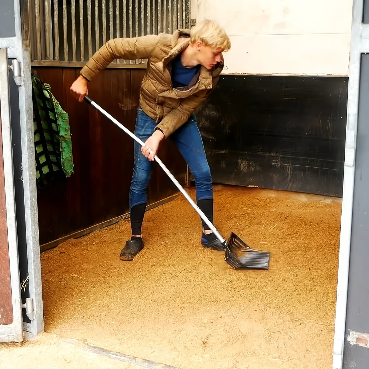 A person in a barn uses a rake to clean the dirt floor, wearing a brown jacket and jeans.