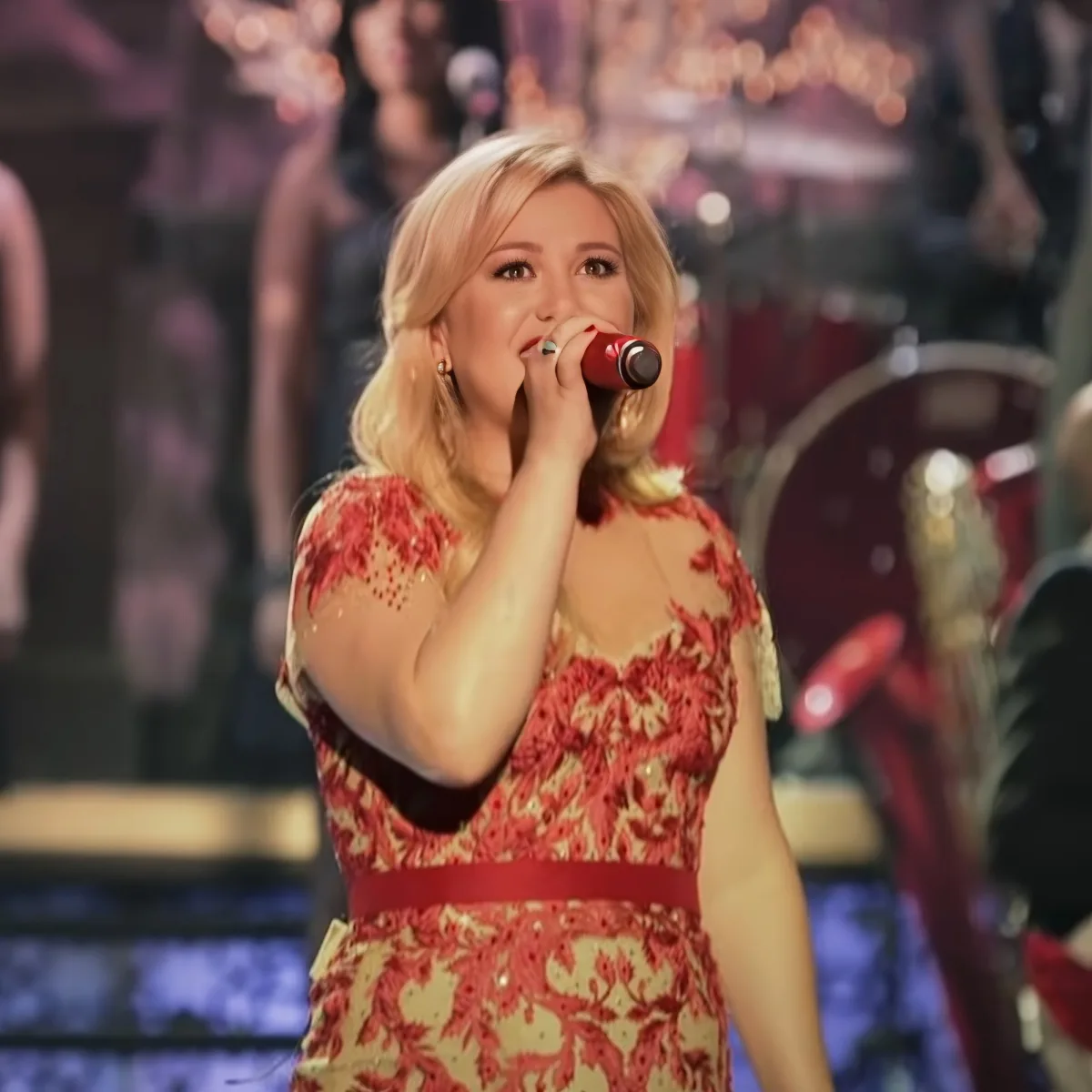 A woman in a red floral dress sings into a microphone on stage.