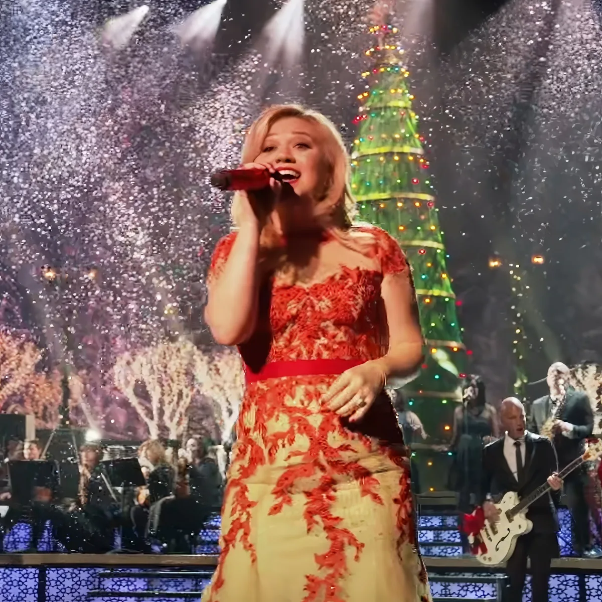 A woman in a red and white dress sings into a microphone on stage with a Christmas tree and confetti in the background.