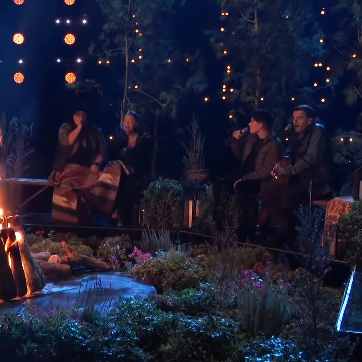 Four people sit around a campfire in a forest setting, illuminated by string lights.