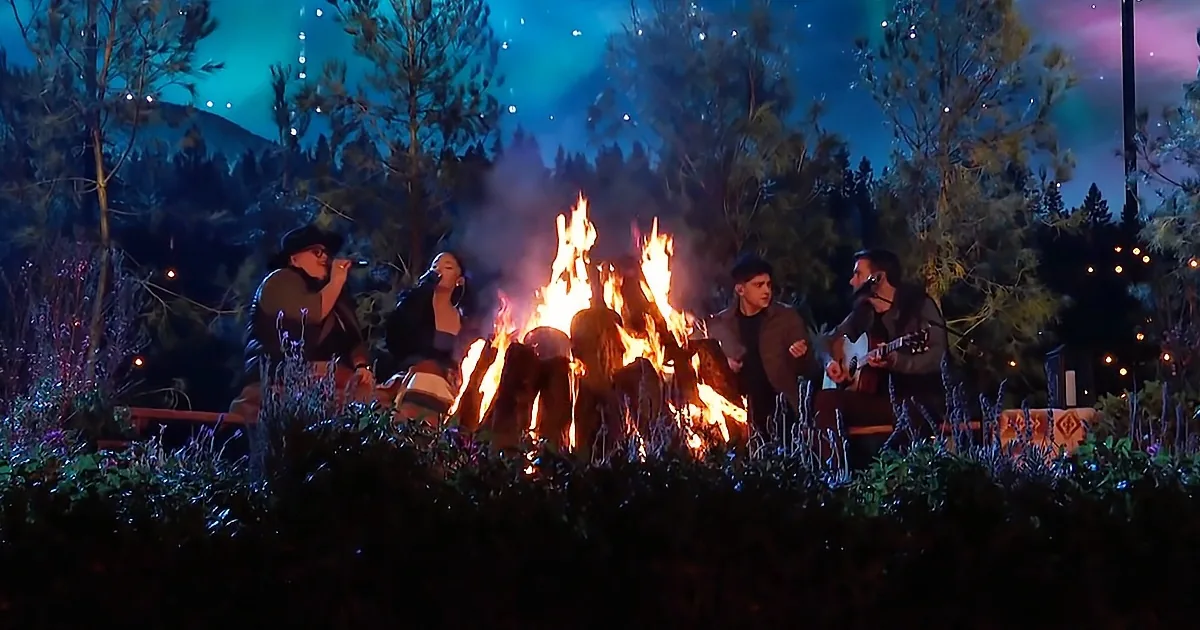 A group of people sing and play guitar around a campfire under a starry night sky.