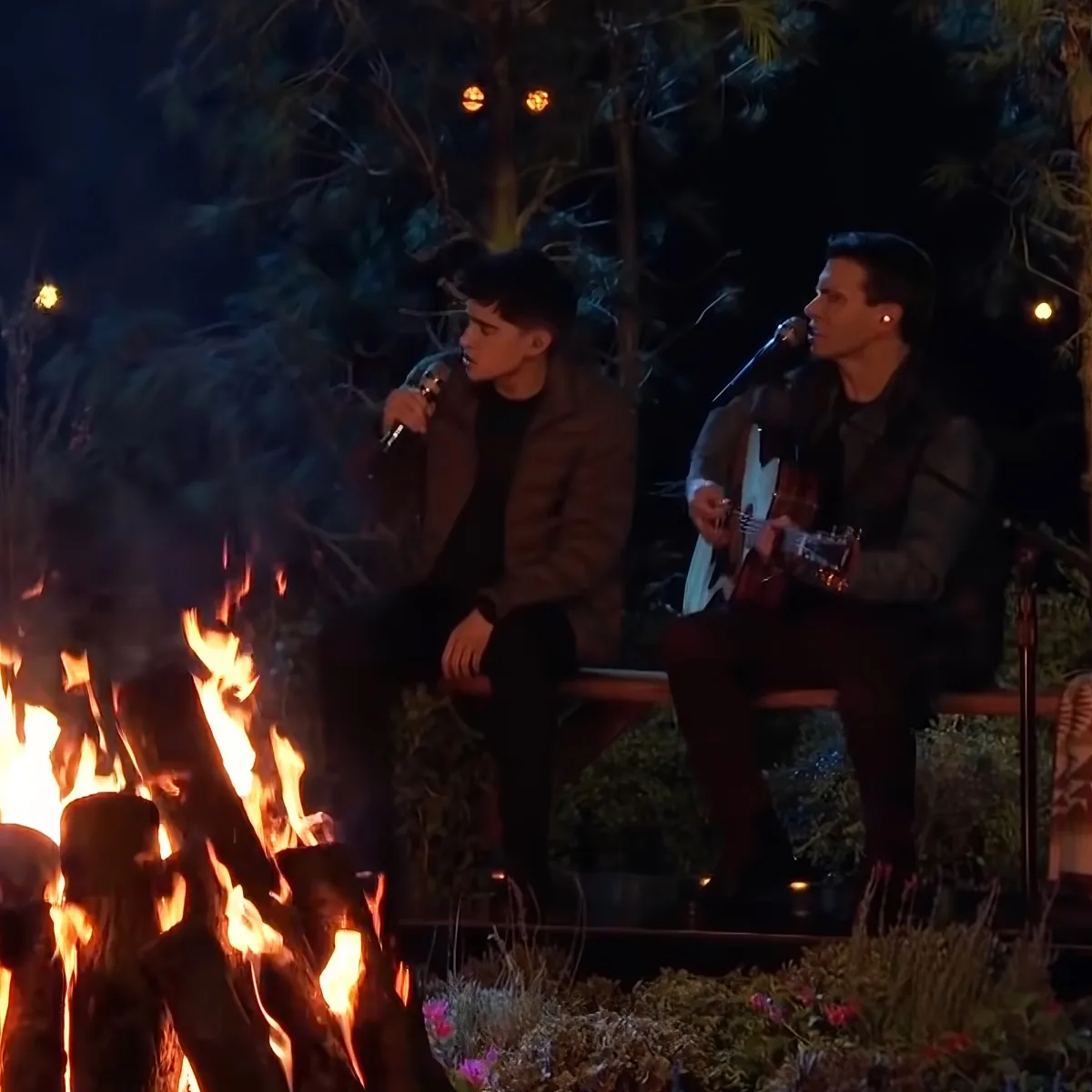 Two men sing by a campfire, one playing guitar, surrounded by trees and lights.