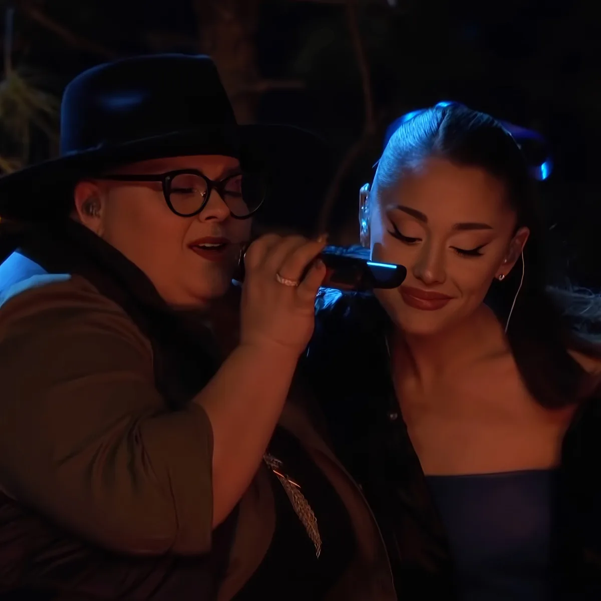 A woman in a hat sings into a microphone while another woman listens nearby.
